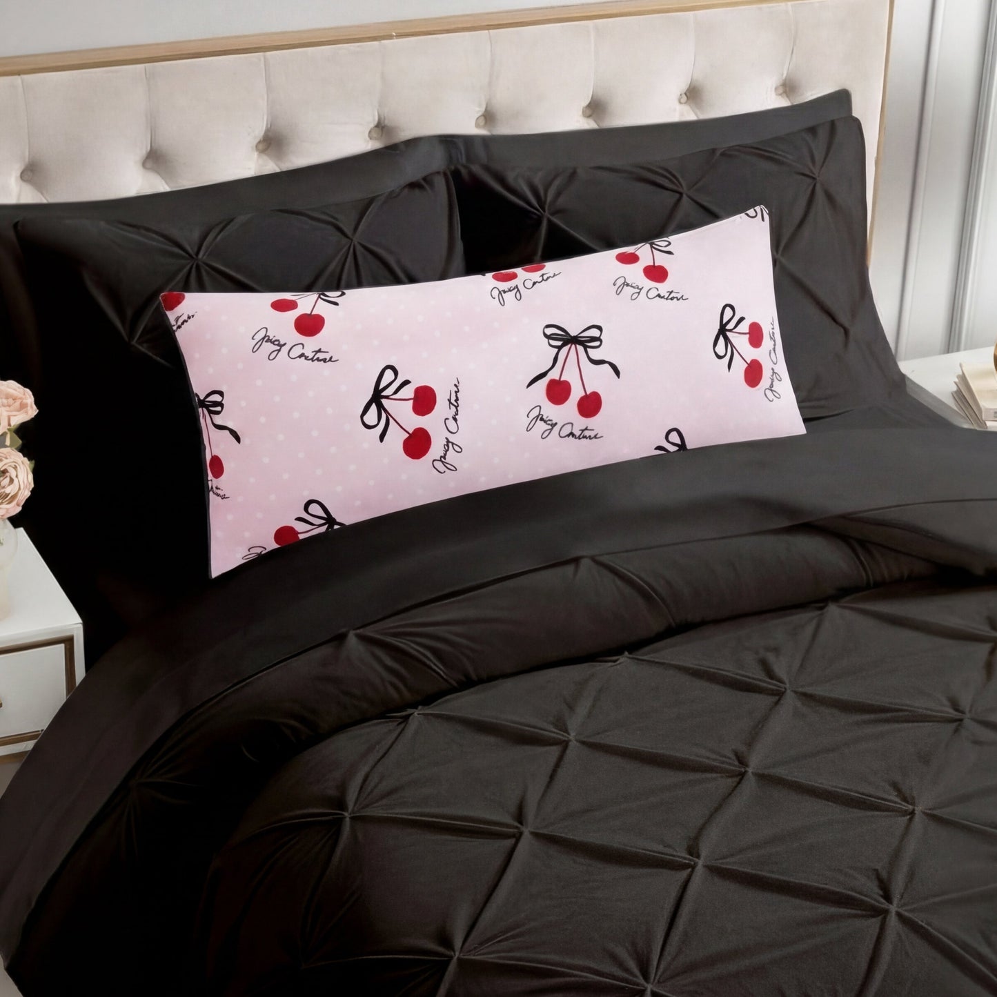 A bed with a black tufted comforter and matching pillowcases features the Plush Body Pillow—oversized, plush, and pink with red cherries, black bows, and cursive accents. The chic pattern pops against the beige tufted headboard.