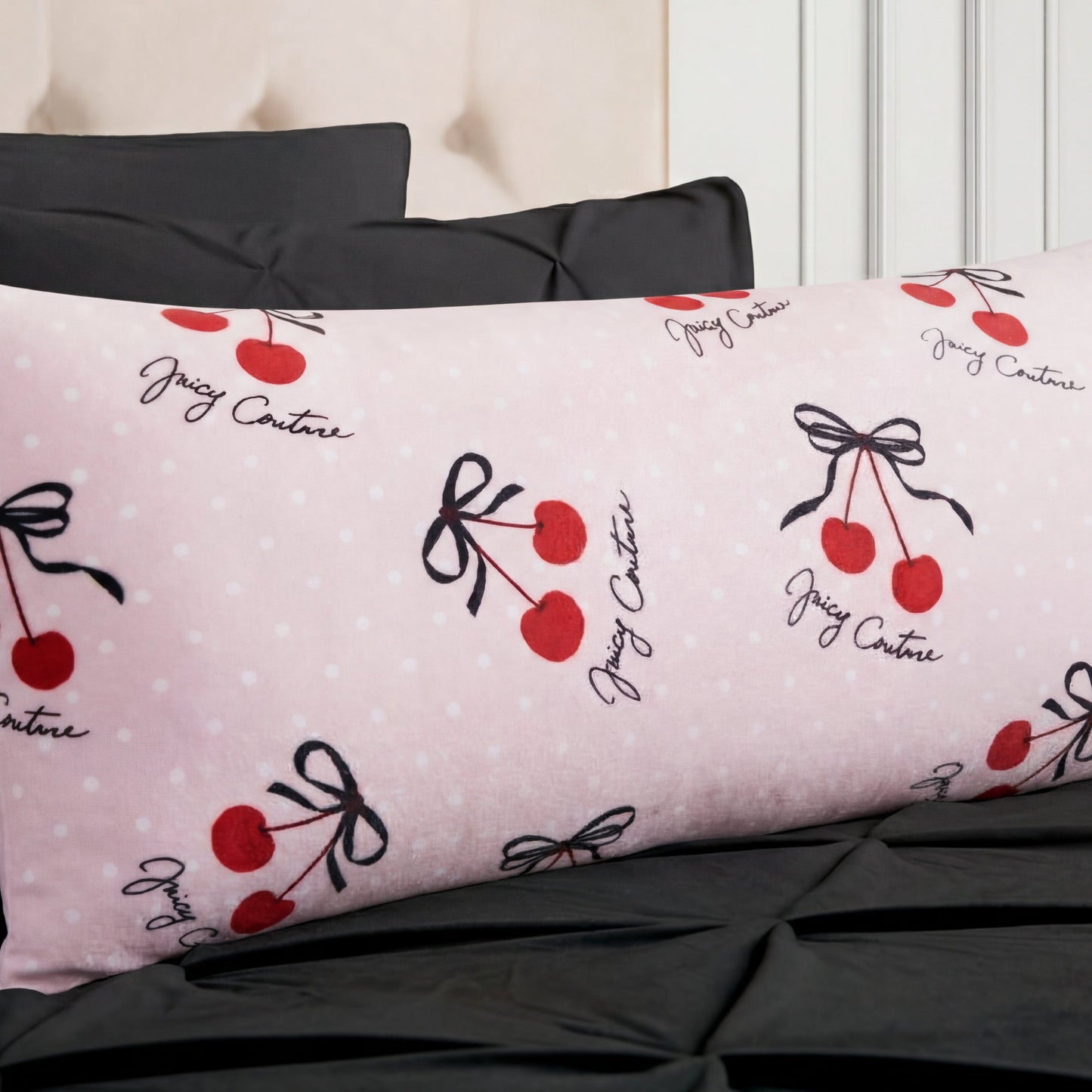The Plush Body Pillow features a light pink Juicy Couture case with white polka dots, red cherries with black bows, and Juicy Couture script in black, shown on a bed with black pillows and bedspread.