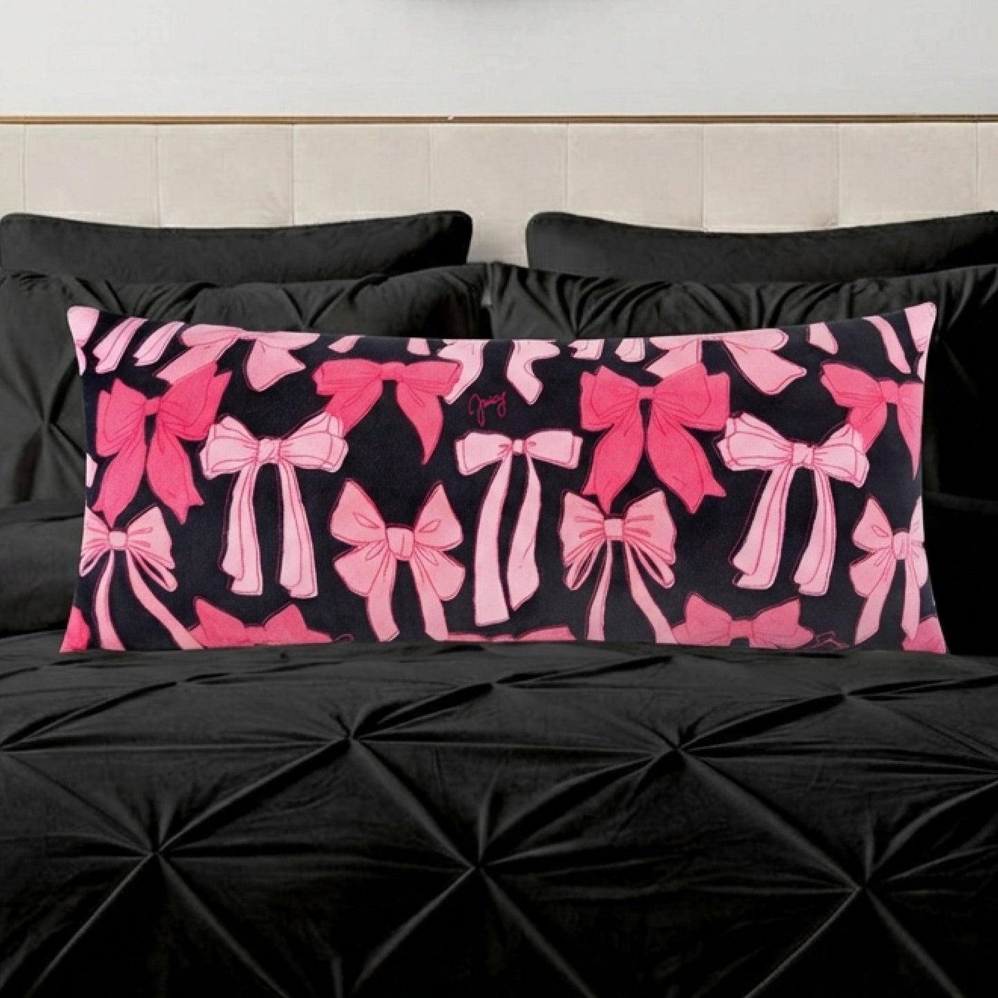 The Plush Body Pillow features an oversized, plush design with various pink bows on a black background, resting on a textured black bedspread with matching pillows.
