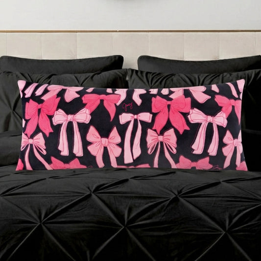 The Plush Body Pillow features an oversized, plush design with various pink bows on a black background, resting on a textured black bedspread with matching pillows.