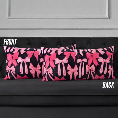 Two rectangular Plush Body Pillows with a pink bow pattern on a dark background are displayed on a black tufted sofa. Labels "FRONT" and "BACK" show both sides feature the same bow design.
