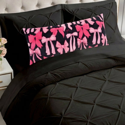 A bed with black, textured bedding and a Plush Body Pillow featuring pink and red bows sits against a light-colored, tufted headboard, next to a nightstand with a vase of flowers.