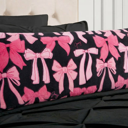 The Plush Body Pillow by Juicy Couture features pink bows and cursive signatures, adding comfort and designer flair to your space. Styled on a bed with black bedding and a beige tufted headboard for a chic look.