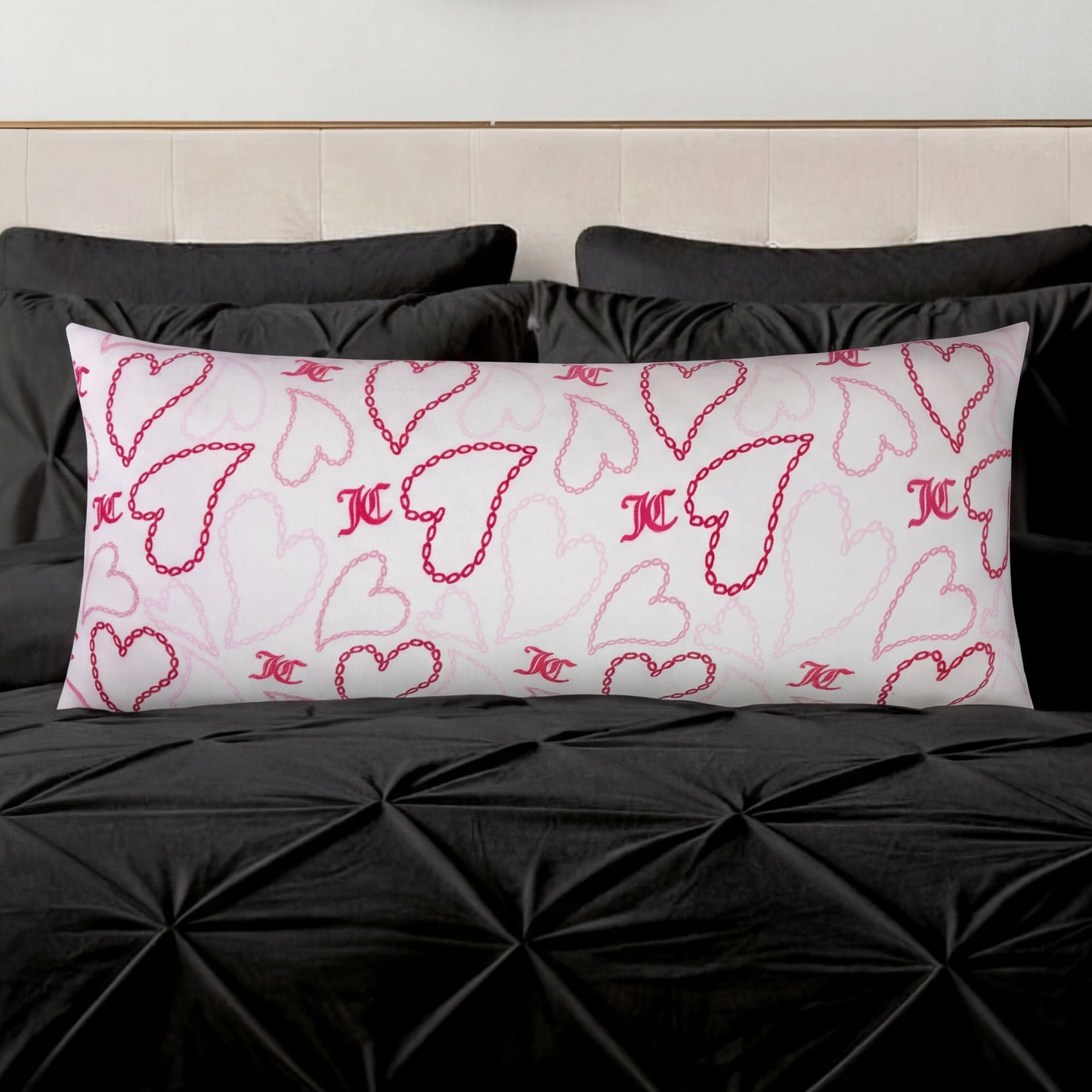 The Plush Body Pillow, featuring pink and red heart designs and decorative initials, rests on a black tufted bedspread with black pillows in the background.