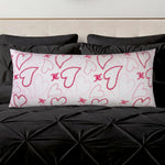 The Plush Body Pillow, featuring pink and red heart designs and decorative initials, rests on a black tufted bedspread with black pillows in the background.