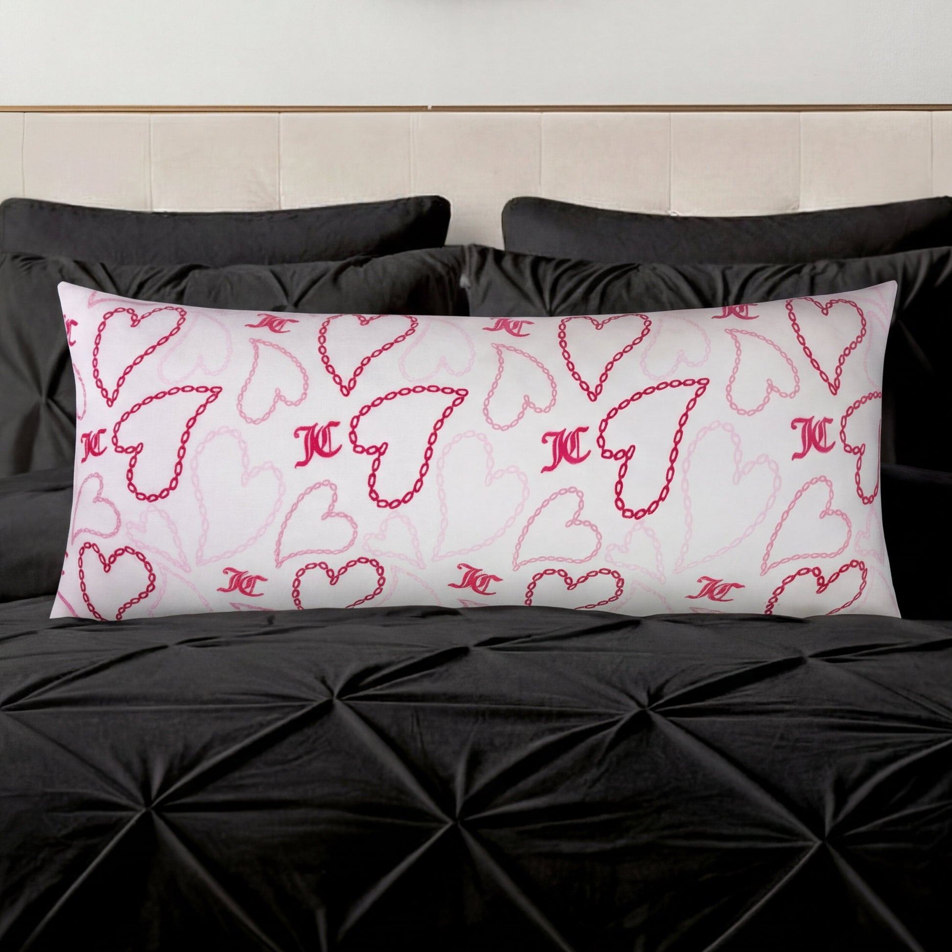 The Plush Body Pillow, featuring pink and red heart designs and decorative initials, rests on a black tufted bedspread with black pillows in the background.