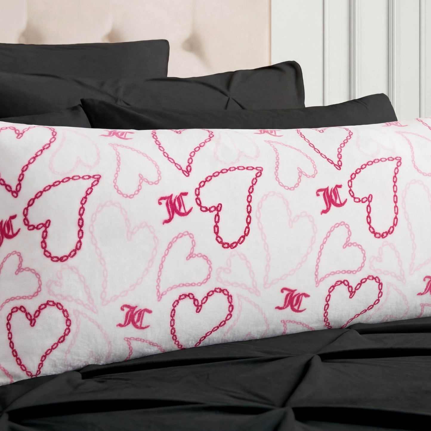 The Plush Body Pillow features pink heart and chain patterns and rests on a black bedspread, paired with black pillows and a beige upholstered headboard.