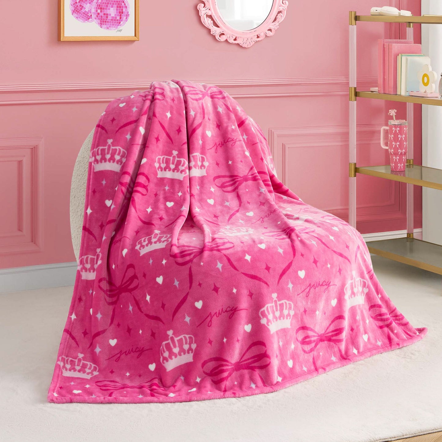 A Kids Plush Throw Blanket in plush pink with white crowns, bows, and “juicy” is draped over a white chair in a chic pink room featuring wall art, a mirror, and stylish shelves.