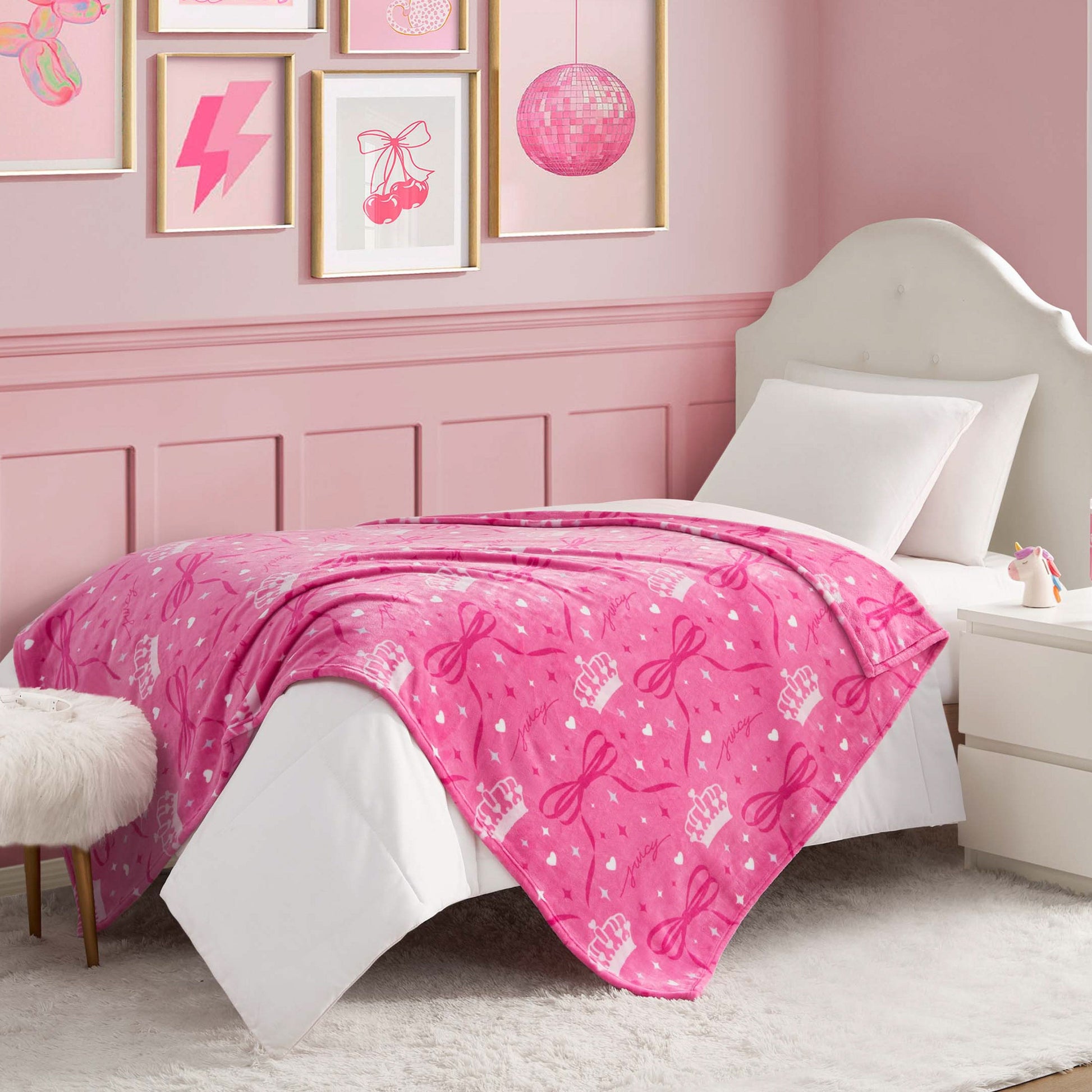 A neatly made bed with a bright pink Kids Plush Throw Blanket featuring bows and birthday cakes, white pillows, and a white headboard. The room has pink walls, framed art, and a white nightstand with a lamp.