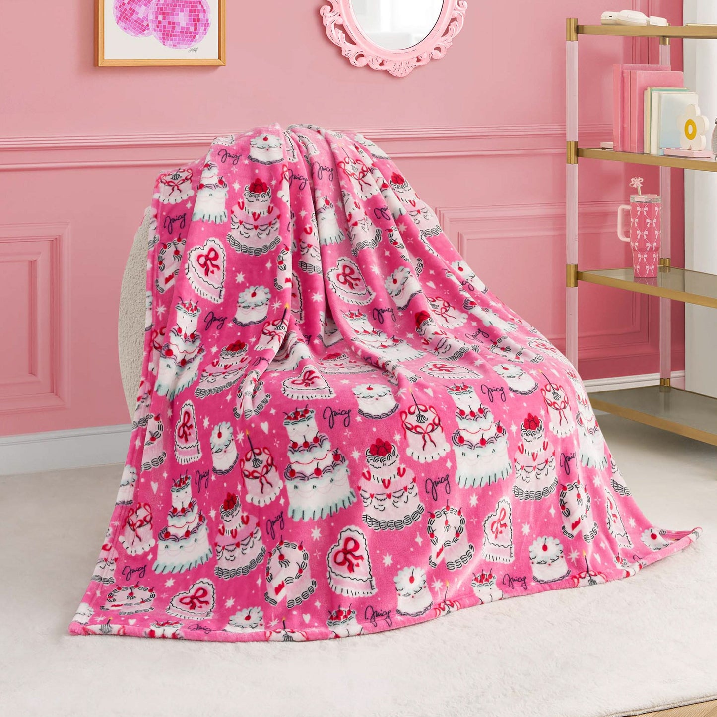 The Kids Plush Throw Blanket, featuring Hello Kitty with cakes, crowns, and bows in pink, is draped over a chair in a girly-themed room with decorative wall art and a shelf in the background.
