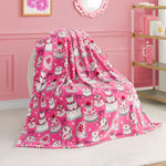 The Kids Plush Throw Blanket, featuring Hello Kitty with cakes, crowns, and bows in pink, is draped over a chair in a girly-themed room with decorative wall art and a shelf in the background.
