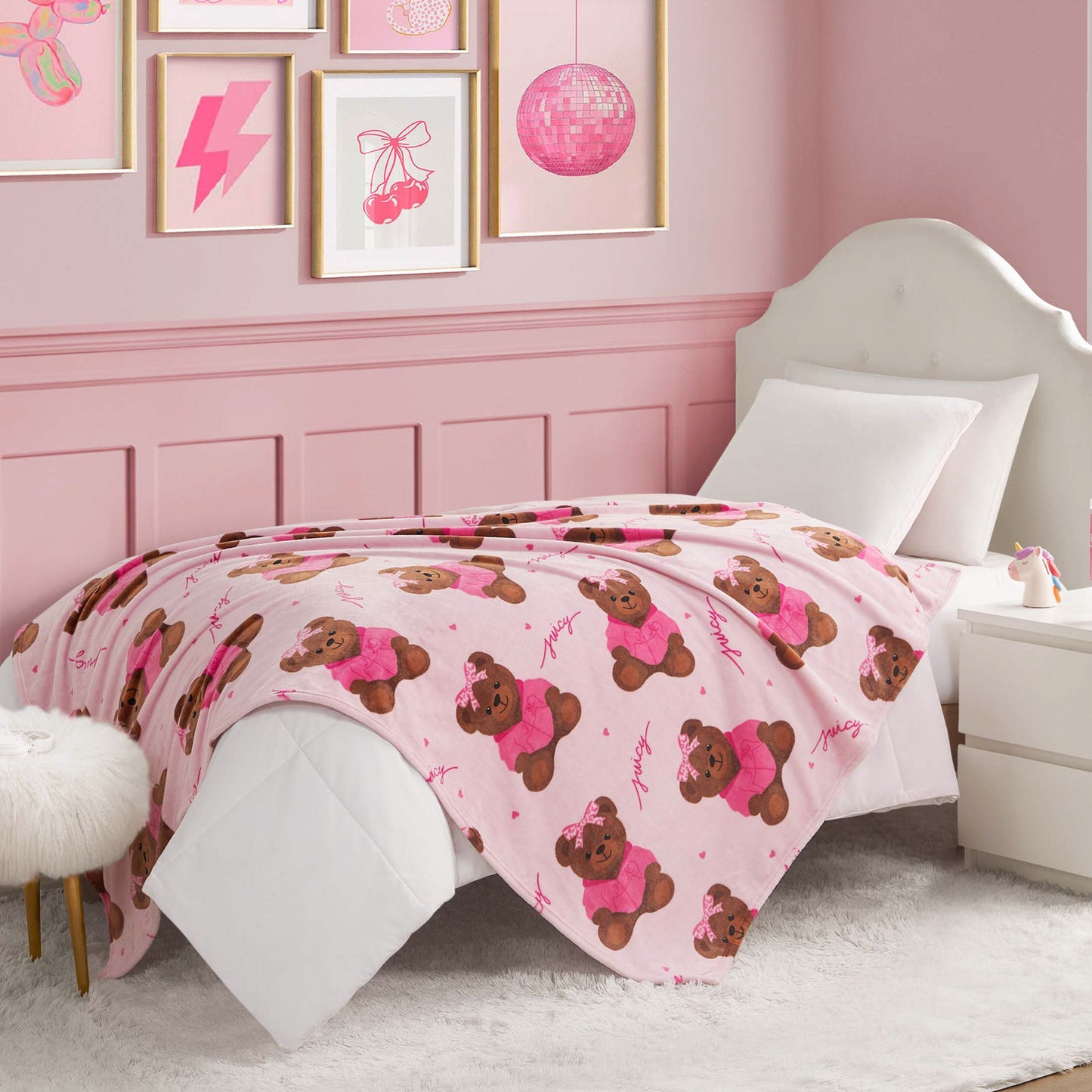 A neatly made bed with a white headboard and a Kids Plush Throw Blanket decorated with brown teddy bears. The room has pink walls, white furniture, and pink-themed artwork above the bed.