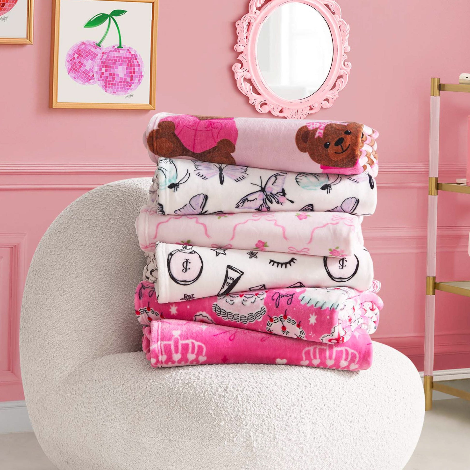 A stack of five ultra-soft blankets, including a Kids Plush Throw Blanket, sits on a white textured chair in a pink room with framed cherry artwork and a round wall mirror.