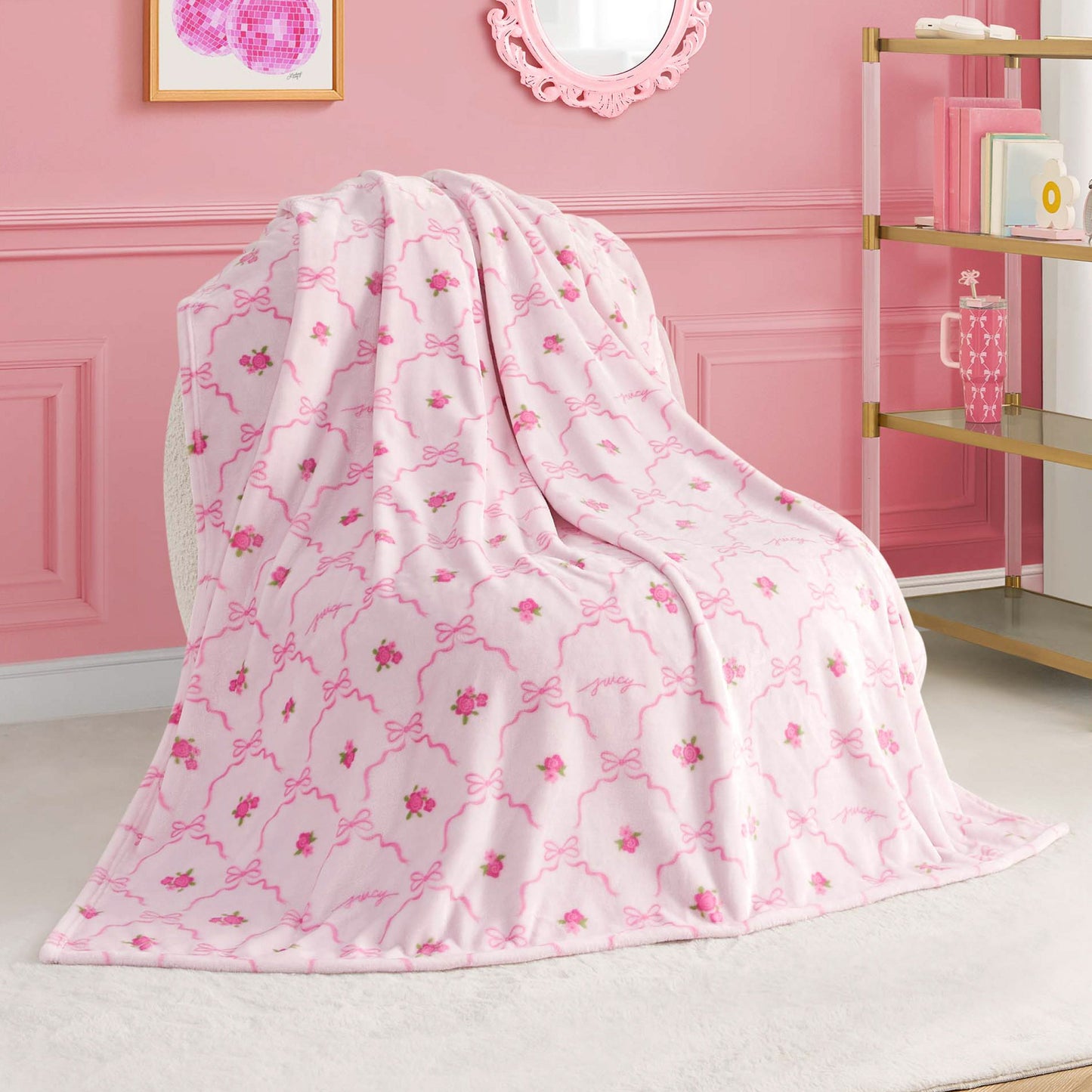 A Kids Plush Throw Blanket from Juicy Couture, ultra-soft in plush pink with floral pattern and decorative lines, is draped over a chair in a pink-themed room featuring gold and white shelves and framed wall art.