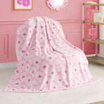 A Kids Plush Throw Blanket from Juicy Couture, ultra-soft in plush pink with floral pattern and decorative lines, is draped over a chair in a pink-themed room featuring gold and white shelves and framed wall art.