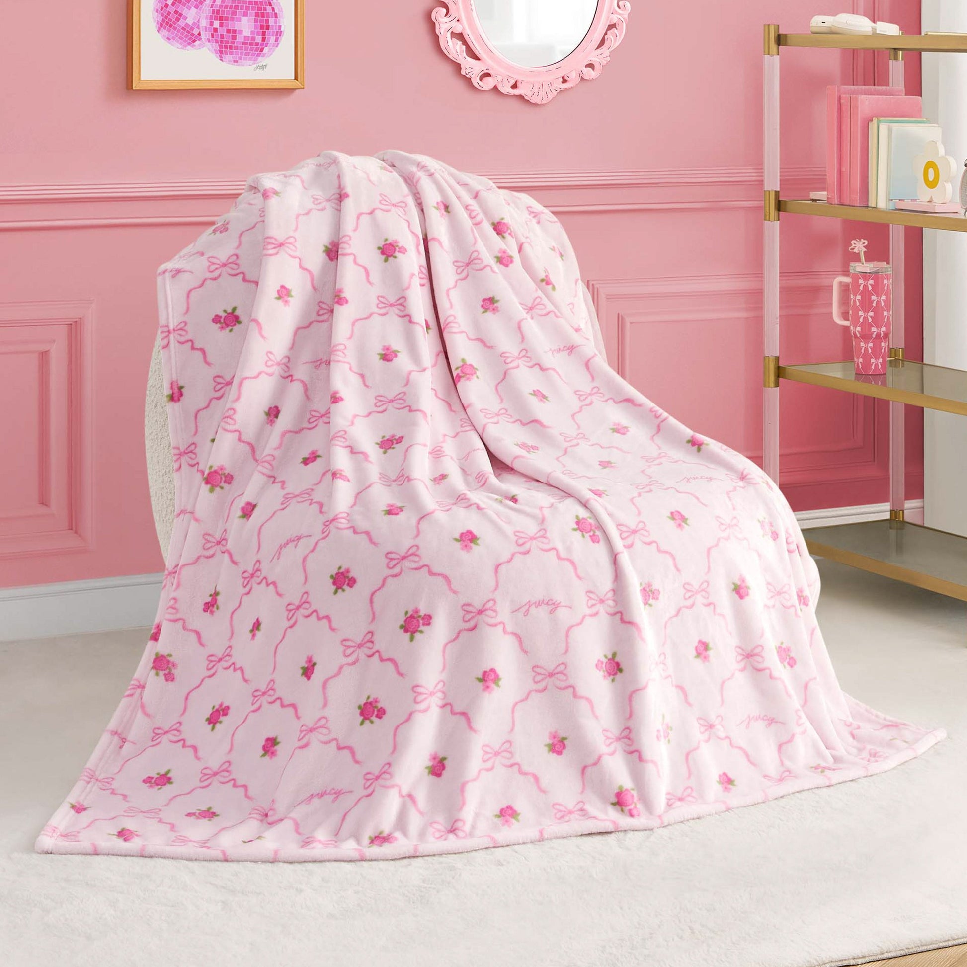 A Kids Plush Throw Blanket from Juicy Couture, ultra-soft in plush pink with floral pattern and decorative lines, is draped over a chair in a pink-themed room featuring gold and white shelves and framed wall art.