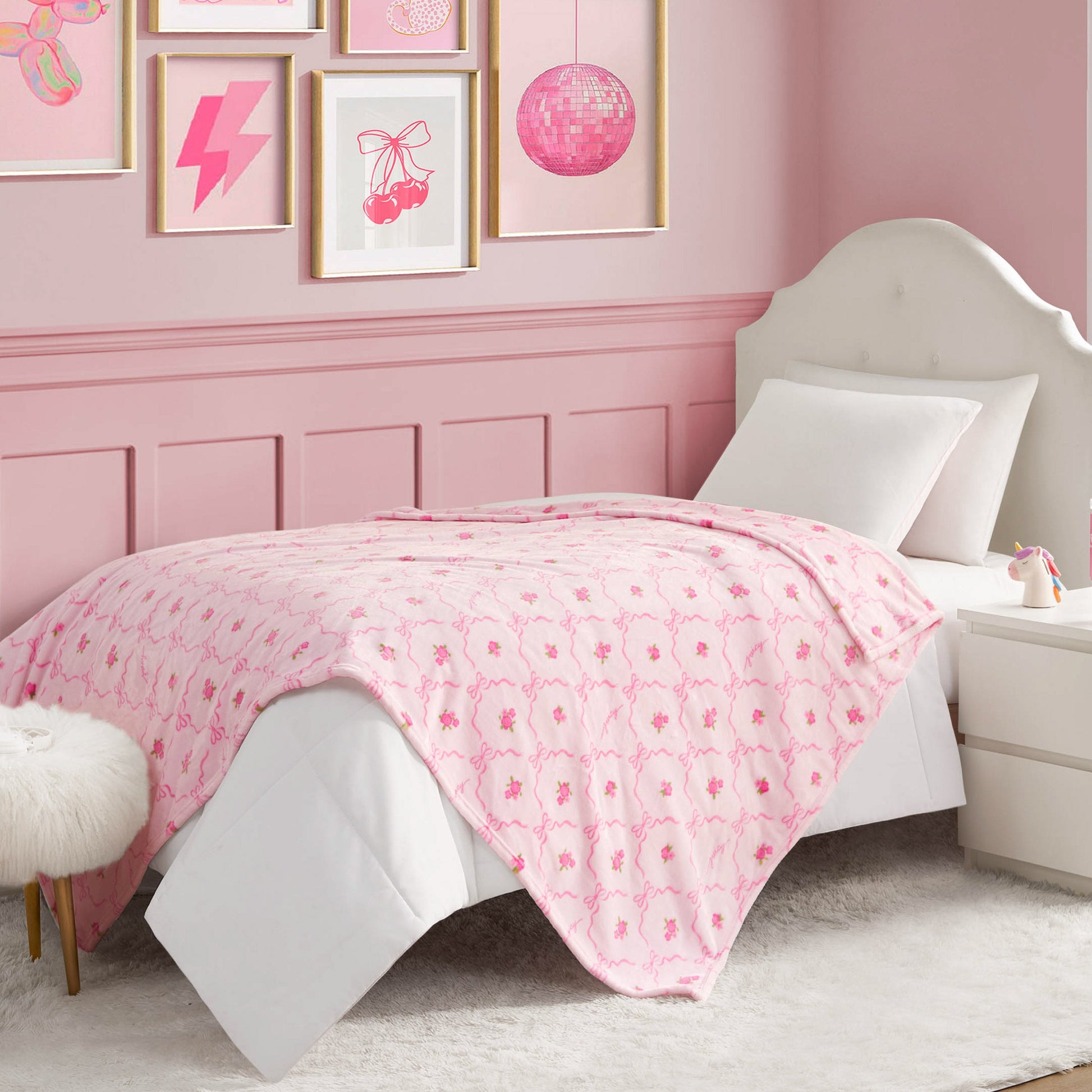 A neatly made bed features a Kids Plush Throw Blanket with a pink and white floral pattern in a pink-themed room, complete with framed art, a white nightstand, and a fuzzy stool for a cozy, stylish vibe.
