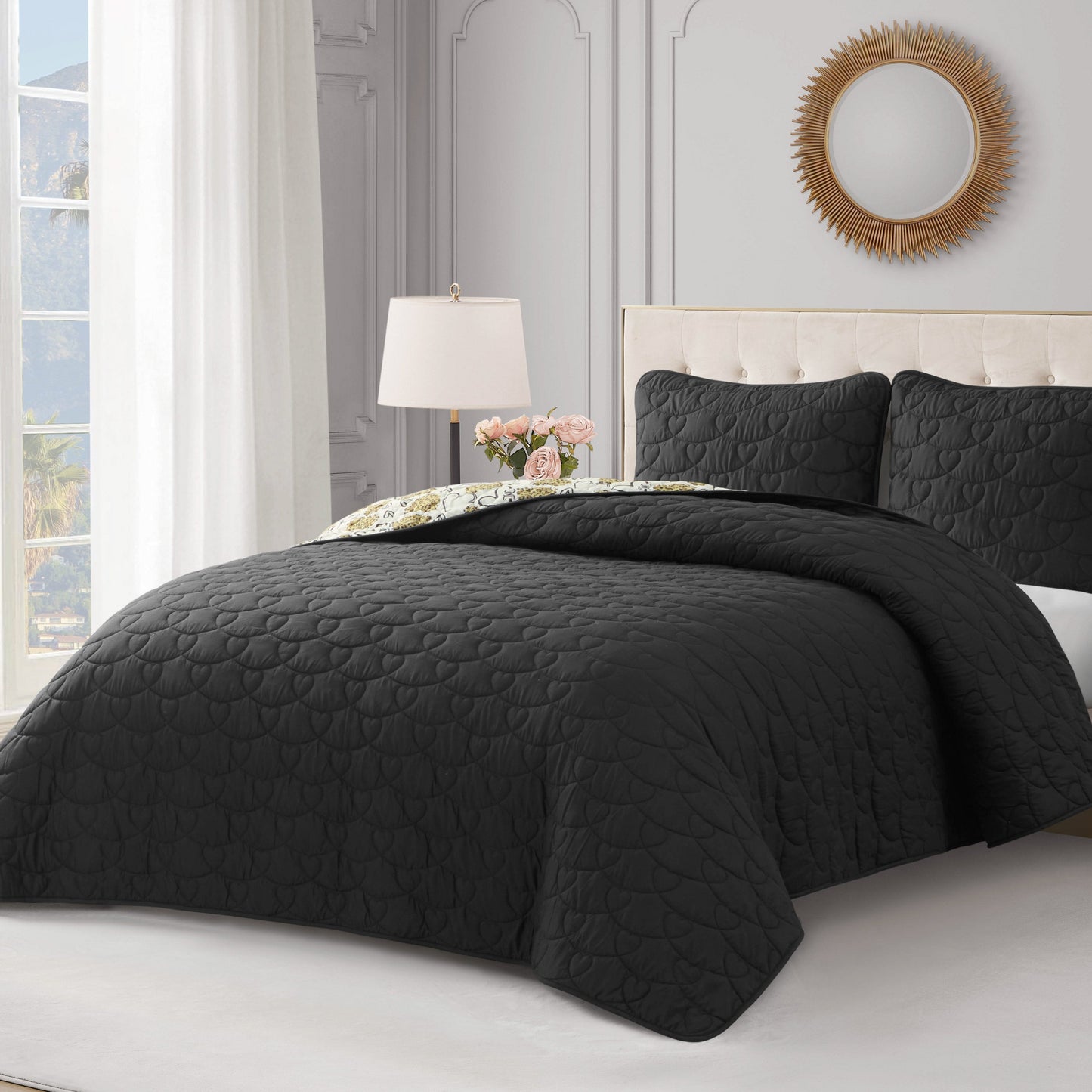 A neatly made bed features the Cheetah Doodle Hearts Quilt Set with a black quilted, reversible quilt and matching pillows in an elegant bedroom with white walls, a sunburst mirror, a bedside lamp, and pink flowers on the nightstand.