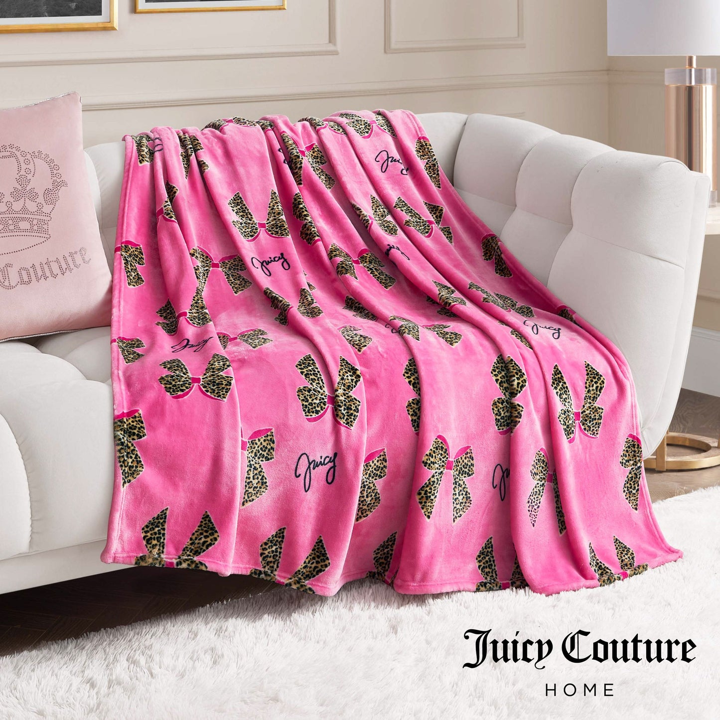 The Plush Oversized Throw Blanket in plush pink, decorated with leopard print bows and Juicy text, is draped over a white sofa and paired with a pink crown pillow for stylish flair in a modern living room.