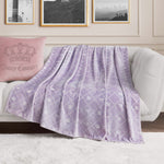The Plush Oversized Throw Blanket in lavender, featuring white crown and Juicy Couture logos, drapes over a white couch beside a pink pillow with matching motifs, adding ultra-soft luxury beneath black-and-white framed art.