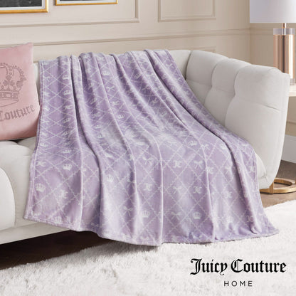 A Plush Oversized Throw Blanket by Juicy Couture, in light purple with a crown and JC logo pattern, drapes over a white sofa beside a pink Juicy Couture pillow. The ultra-soft blanket adds comfort and style. "Juicy Couture Home" is at the bottom right.