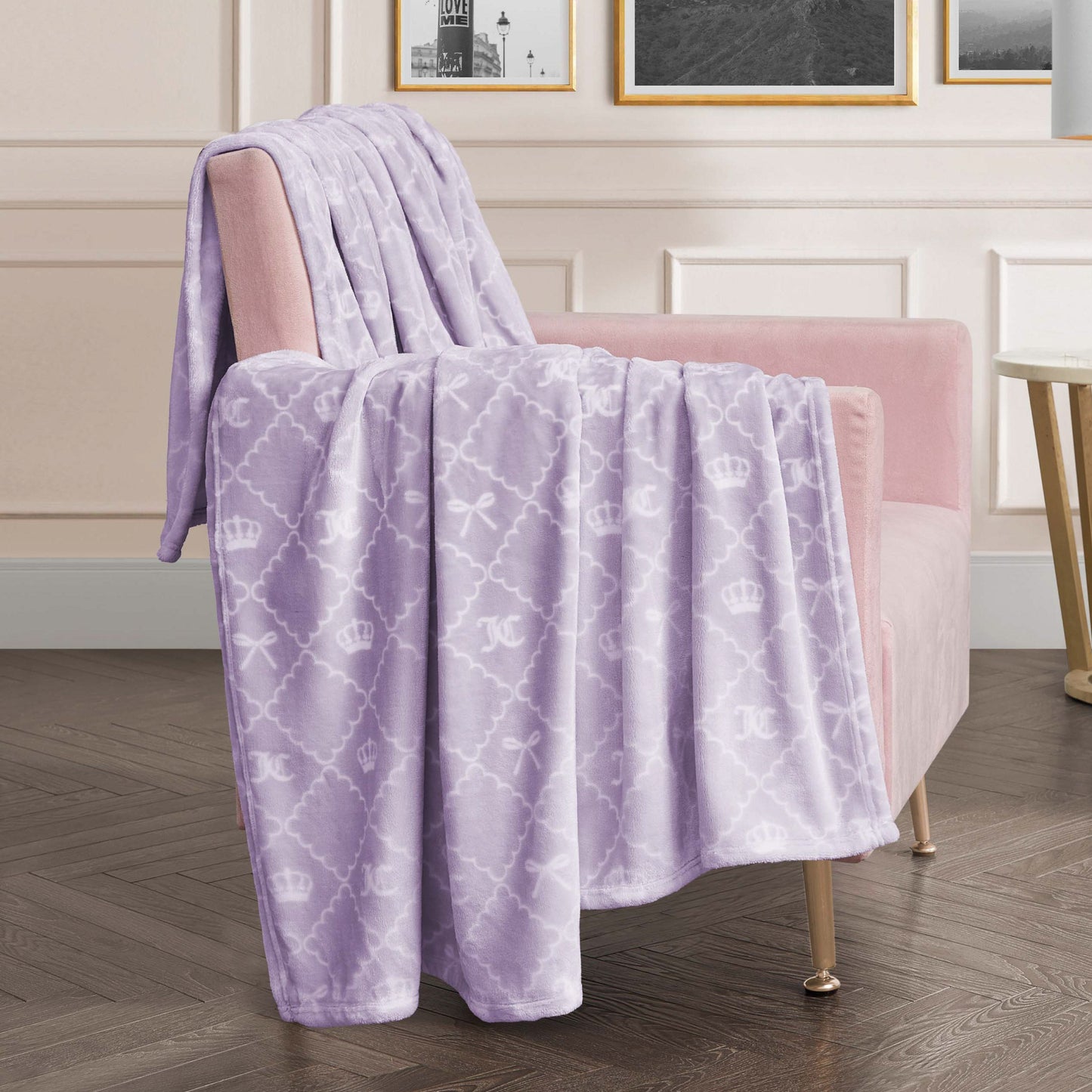 A Plush Oversized Throw Blanket in soft lavender with a white crown and bow design is draped over a blush pink armchair in a modern room with framed art and wood flooring.
