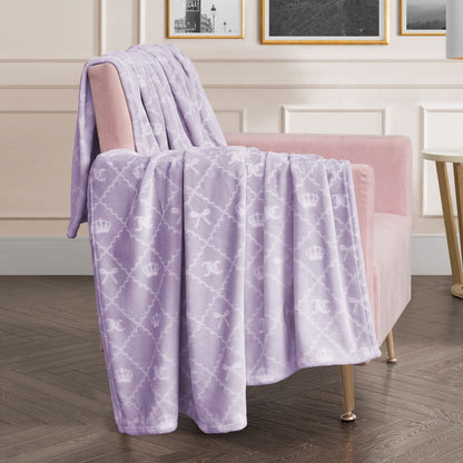 A Plush Oversized Throw Blanket in soft lavender with a white crown and bow design is draped over a blush pink armchair in a modern room with framed art and wood flooring.