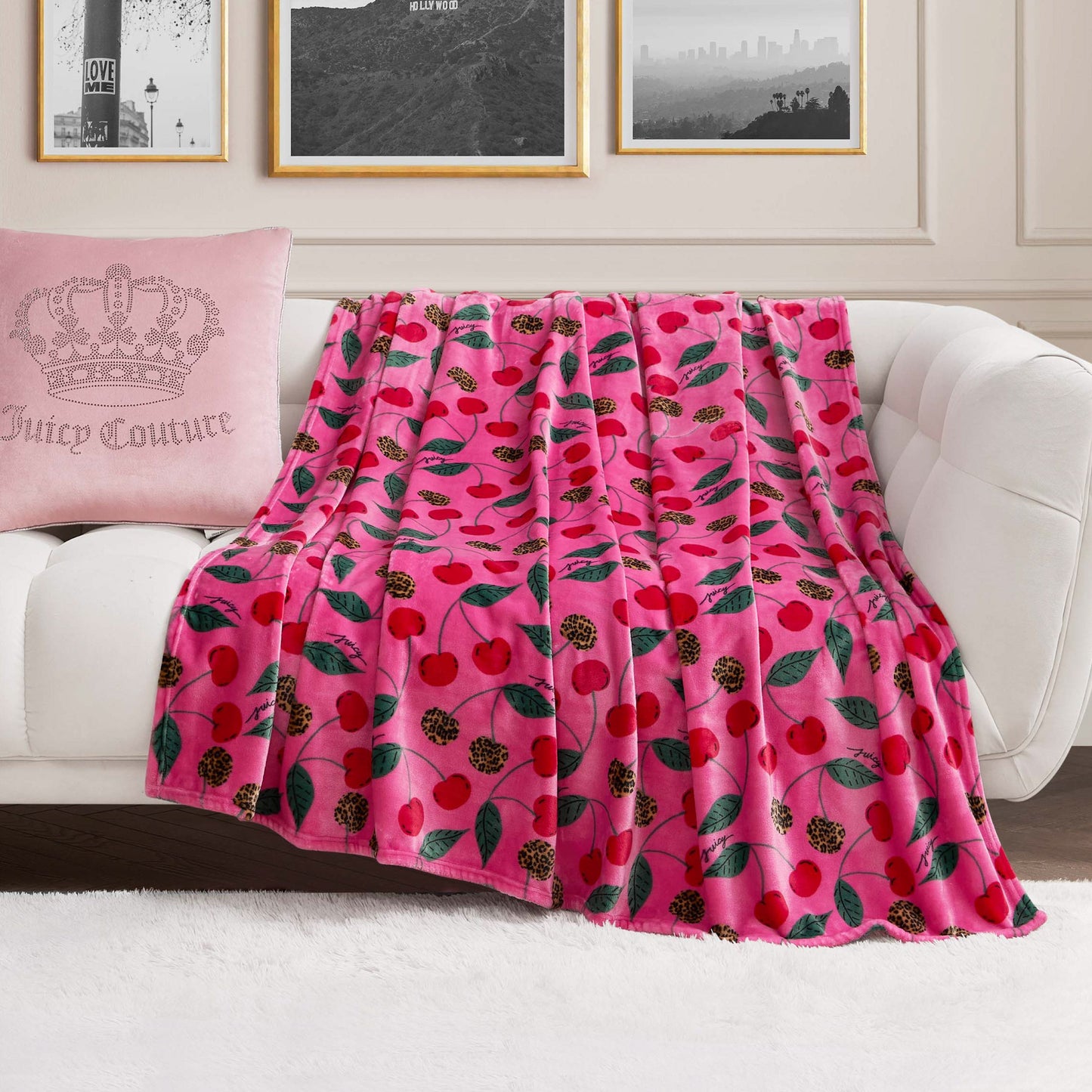 A Plush Oversized Throw Blanket in pink with a cherry and leaf print is draped over a white sofa. At one end sits a pale pink crown pillow with Juicy Couture text, set against black-and-white photos on the wall behind.
