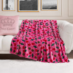 A Plush Oversized Throw Blanket in pink with a cherry and leaf print is draped over a white sofa. At one end sits a pale pink crown pillow with Juicy Couture text, set against black-and-white photos on the wall behind.