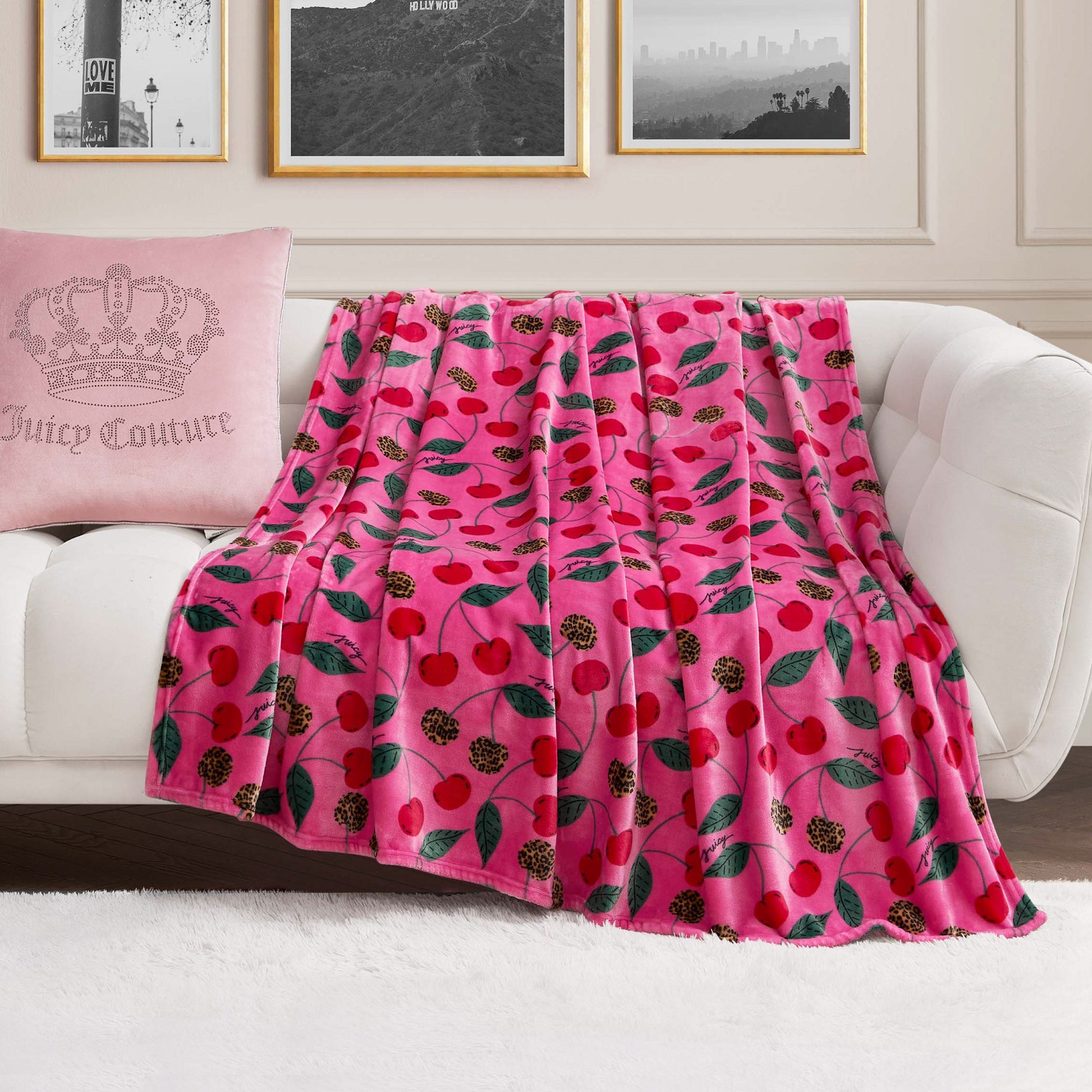 A Plush Oversized Throw Blanket in pink with a cherry and leaf print is draped over a white sofa. At one end sits a pale pink crown pillow with Juicy Couture text, set against black-and-white photos on the wall behind.