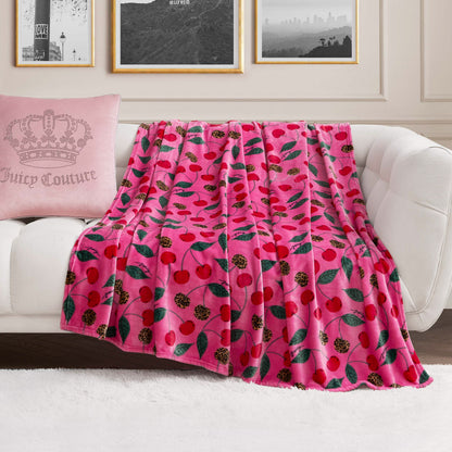 A Plush Oversized Throw Blanket in pink with a cherry and leaf print is draped over a white sofa. At one end sits a pale pink crown pillow with Juicy Couture text, set against black-and-white photos on the wall behind.