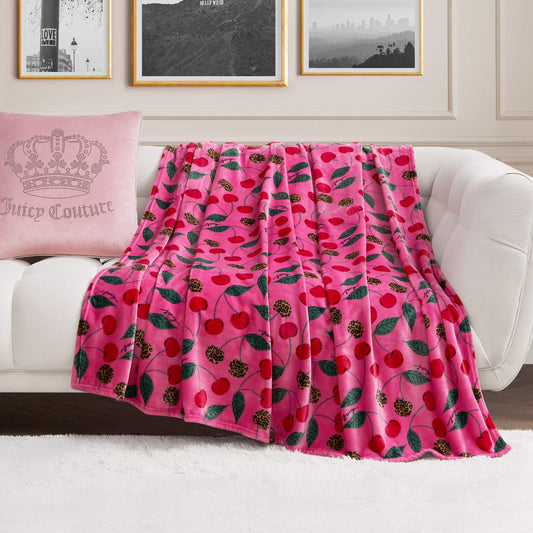 A Plush Oversized Throw Blanket in pink with a cherry and leaf print is draped over a white sofa. At one end sits a pale pink crown pillow with Juicy Couture text, set against black-and-white photos on the wall behind.