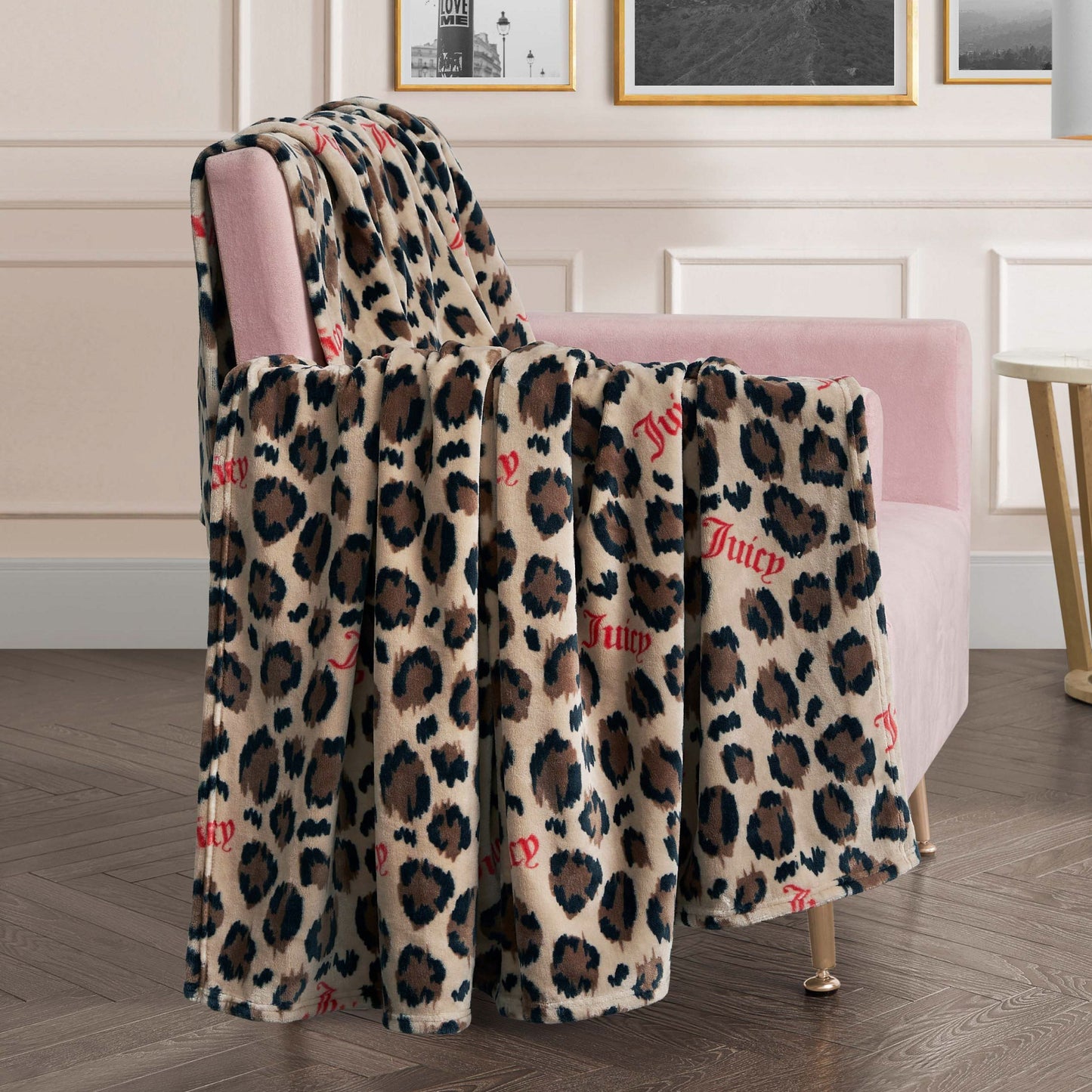 A Plush Oversized Throw Blanket in beige and brown, featuring "Juicy" in red script, is draped over a pink velvet chair in a chic living room with framed art on the wall.