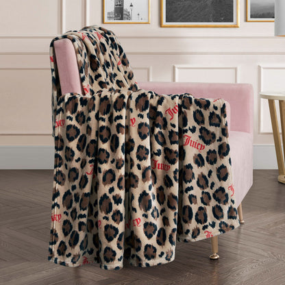 A Plush Oversized Throw Blanket in beige and brown, featuring "Juicy" in red script, is draped over a pink velvet chair in a chic living room with framed art on the wall.