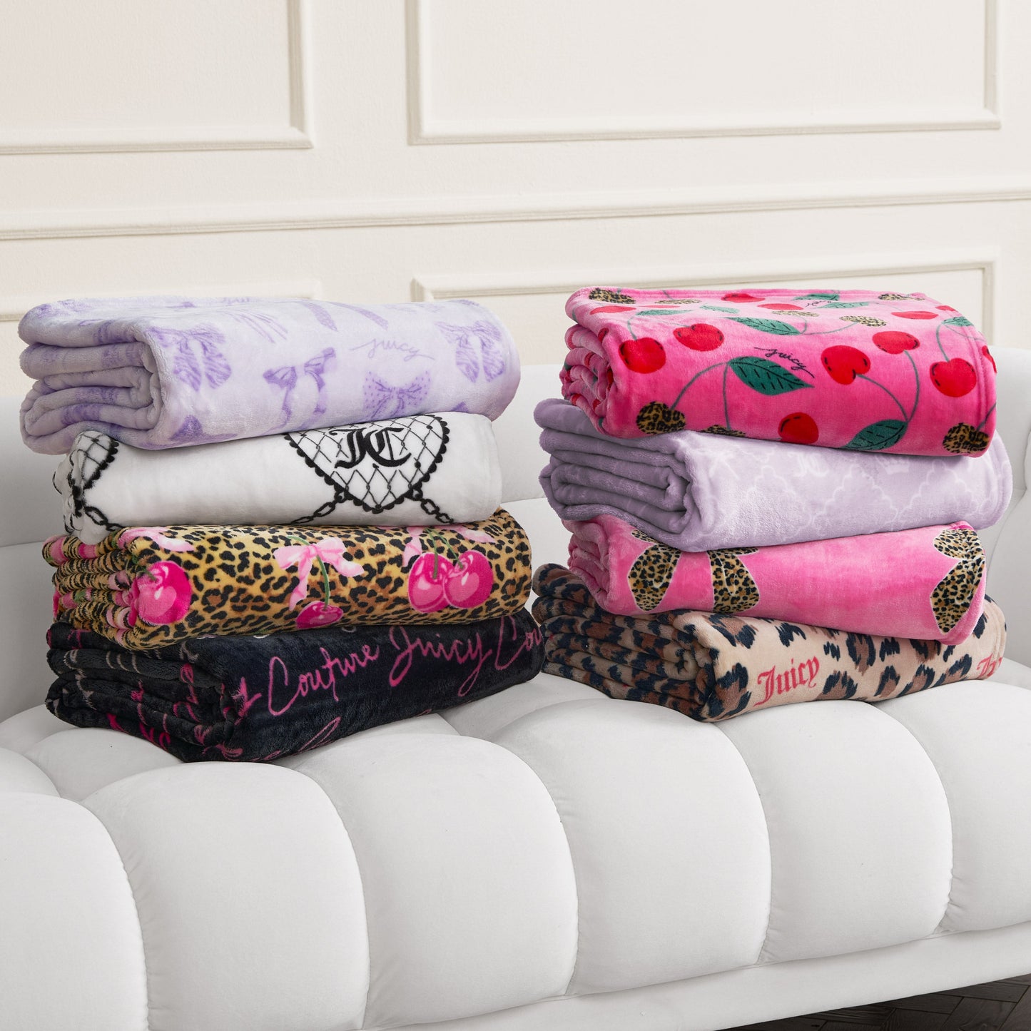 Six folded blankets, including a Plush Oversized Throw Blanket, in various colors and patterns—purple, white, pink, cherry print, leopard print—stacked on a white tufted bench against a paneled wall.
