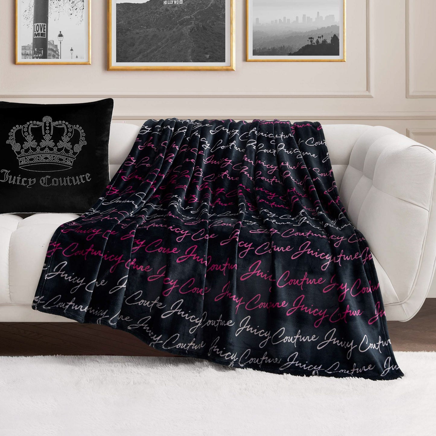 A Plush Oversized Throw Blanket with “Juicy Couture” in white and pink script is draped over a white sofa next to a black Juicy Couture pillow, set in a modern living room with framed wall art.