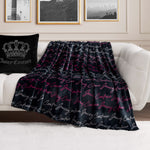 A Plush Oversized Throw Blanket with “Juicy Couture” in white and pink script is draped over a white sofa next to a black Juicy Couture pillow, set in a modern living room with framed wall art.