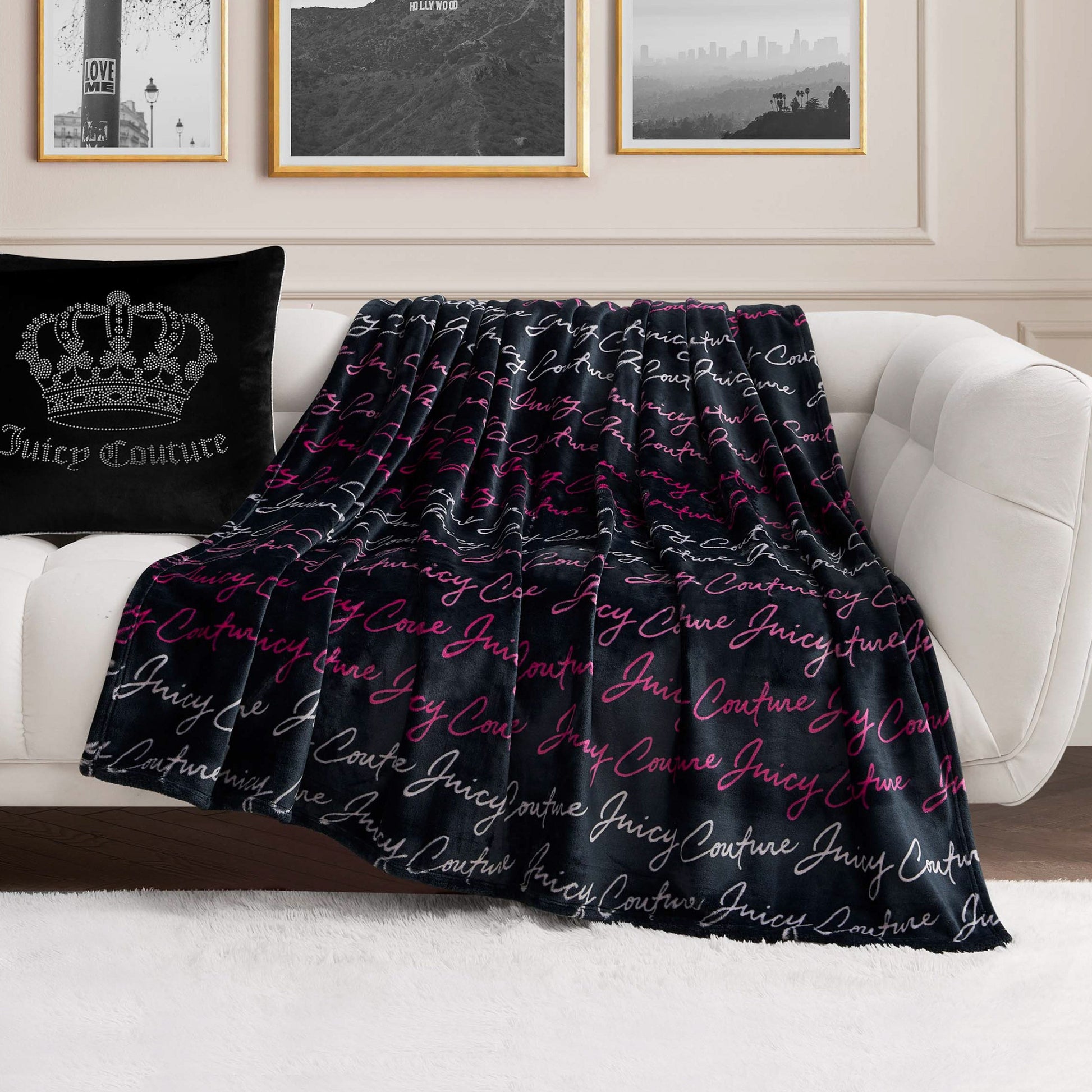 A Plush Oversized Throw Blanket with “Juicy Couture” in white and pink script is draped over a white sofa next to a black Juicy Couture pillow, set in a modern living room with framed wall art.