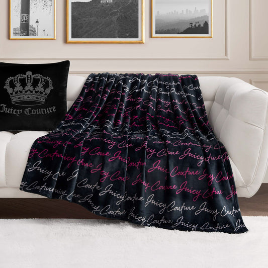 A Plush Oversized Throw Blanket with “Juicy Couture” in white and pink script is draped over a white sofa next to a black Juicy Couture pillow, set in a modern living room with framed wall art.