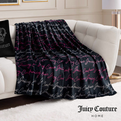 A Plush Oversized Throw Blanket in black with pink and white cursive accents is draped over a white couch, paired with a matching black pillow for a cozy, stylish living room look.