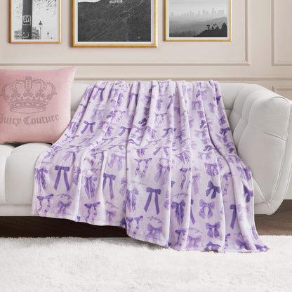 A white couch with a pink Juicy Couture pillow and a Plush Oversized Throw Blanket, light purple with bows and butterflies, draped over it. Behind the couch, framed black-and-white photos are displayed on the wall.