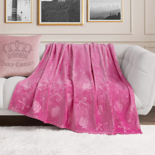 An Embossed Plush Oversized Throw Blanket in bright pink with heart and bow patterns drapes over a white sofa, next to a matching pink pillow with a crown motif, in a chic living room adorned with framed black-and-white photos.