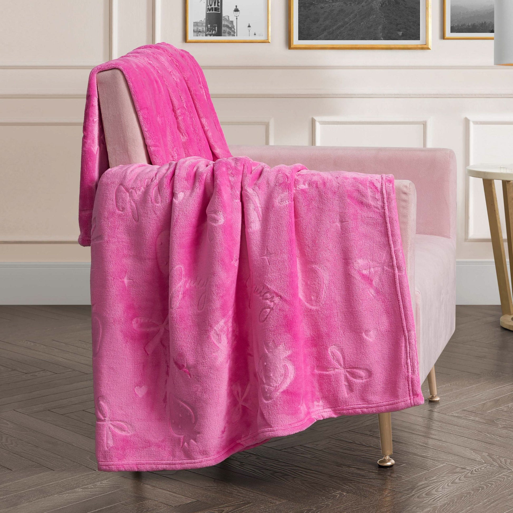 An Embossed Plush Oversized Throw Blanket in soft, bright pink with ribbon and heart designs drapes over a light pink armchair in a modern living room featuring white paneled walls, framed art, and a dark wood floor.