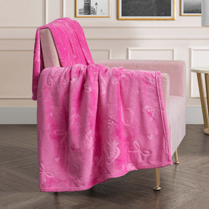 An Embossed Plush Oversized Throw Blanket in soft, bright pink with ribbon and heart designs drapes over a light pink armchair in a modern living room featuring white paneled walls, framed art, and a dark wood floor.