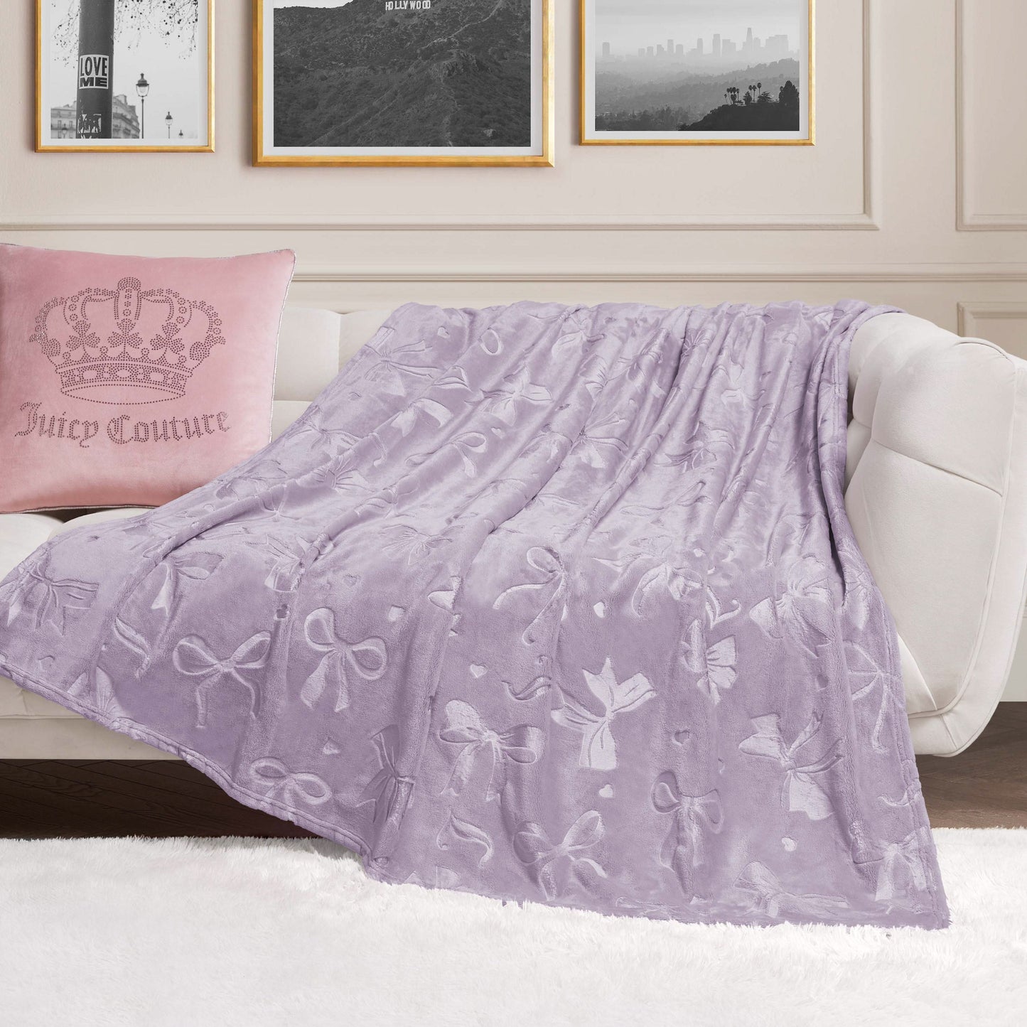 An Embossed Plush Oversized Throw Blanket with soft lavender bow patterns is draped over a white sofa beside a pink Juicy Couture pillow, with black-and-white photos framed on the cream-colored wall behind.