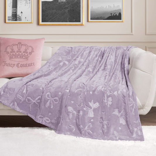 An Embossed Plush Oversized Throw Blanket with soft lavender bow patterns is draped over a white sofa beside a pink Juicy Couture pillow, with black-and-white photos framed on the cream-colored wall behind.