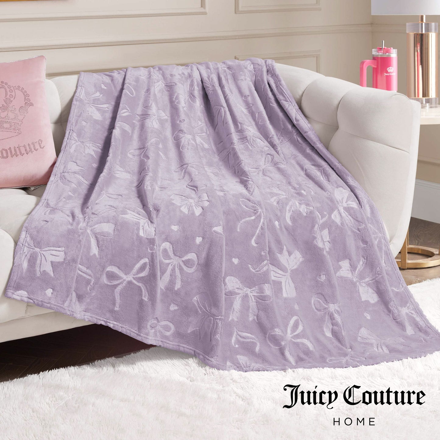 An Embossed Plush Oversized Throw Blanket in light purple with a bow pattern is draped on a white sofa. Nearby are a pink Juicy Couture pillow and tumbler. The Juicy Couture Home logo appears at the bottom right.
