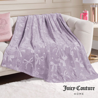 An Embossed Plush Oversized Throw Blanket in light purple with a bow pattern is draped on a white sofa. Nearby are a pink Juicy Couture pillow and tumbler. The Juicy Couture Home logo appears at the bottom right.