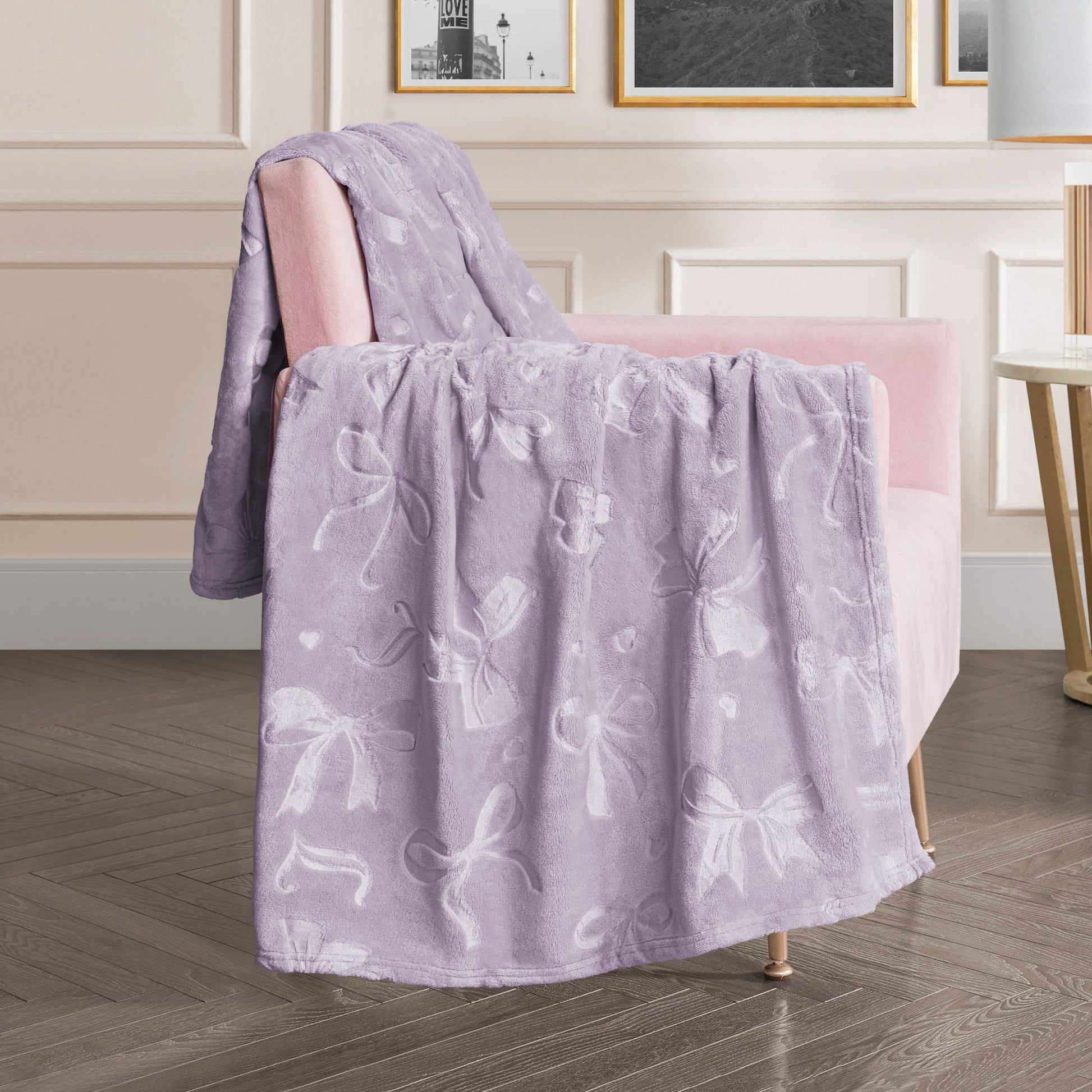 An Embossed Plush Oversized Throw Blanket in light purple rests on a pale pink armchair in a stylish living room featuring soft lighting, framed wall art, and elegant decor.