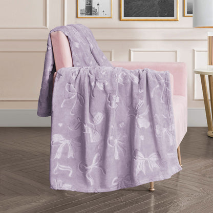 An Embossed Plush Oversized Throw Blanket in light purple rests on a pale pink armchair in a stylish living room featuring soft lighting, framed wall art, and elegant decor.